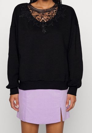 Schwarzer Pullover mit Spitzenausschnitt, langen Ärmeln und gerippten Bündchen, kombiniert mit einem helllila Rock mit seitlichen Schlitzen.