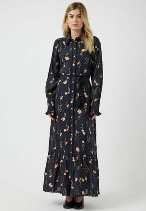 Vrouw draagt een lange zwarte bloemenjurk met knopen, een taillekoord en lange mouwen, staand tegen een effen lichte achtergrond.