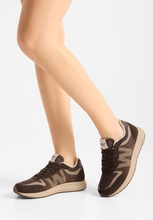 Benen met bruine en beige vetersneakers met een getextureerde zool, geposeerd in een wandelhouding tegen een effen witte achtergrond.