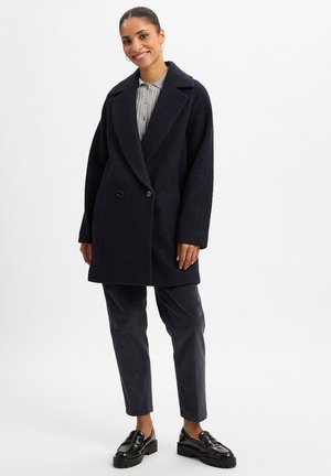 Vrouw die lacht, gekleed in een zwart oversized jas over een grijs geribd shirt, zwarte broek en zwarte glanzende loafers, staand tegen een witte achtergrond.