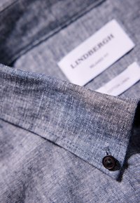 Camisa de mezcla de lino con una tela texturizada de color azul oscuro, cuello y un detalle de botones. Etiquetas visibles en blanco que muestran la información de la marca y del tejido.
