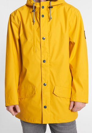 Jasnożółta przeciwdeszczowa kurtka z kapturem, wyposażona w czarne zatrzaski, ściągacze, kieszeń na piersi oraz dwie kieszenie z patkami, noszona przez osobę.