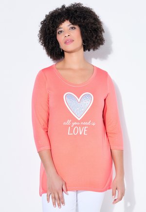 Kobieta o kręconych włosach w koralowej bluzce z rękawami do łokcia, z srebrnym sercem i napisem "wszystko czego potrzebujesz to MIŁOŚĆ", stojąca na białym tle.