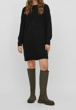 Femme portant une robe-pull noire à manches longues et des bottes vert foncé montant jusqu'aux genoux, debout devant un fond gris clair uni.
