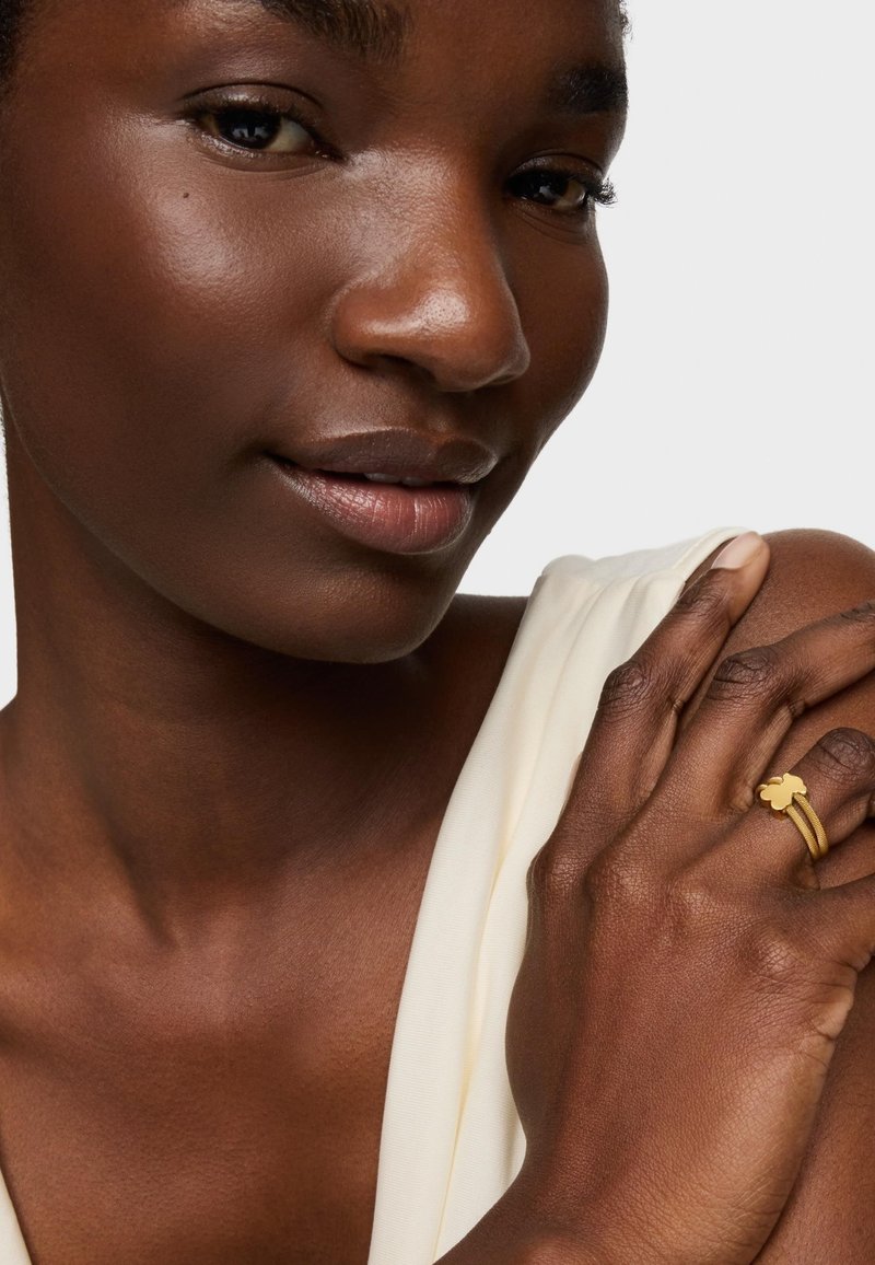 Gold ring featuring a clover design, stacked with two thin bands. Model's skin tone is deep, wearing a light cream-colored garment.