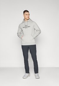 Un sweat à capuche gris avec un logo sur la poitrine, une capuche à cordon, des poches kangourou, accompagné d'un pantalon sombre et de baskets claires, présenté sur un fond uni.