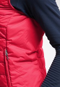 Punainen tilkkupuvun liivi, jossa on sivuvetoketju, käytetty tummansinisen pitkähihaisen paidan päällä. Kyynärvarsi on taipunut kyynärpäätä vasten valkoista taustaa vasten.