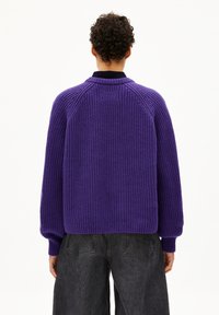 Lila gestrickter Pullover mit strukturiertem Muster, Raglanärmeln und geripptem Saum. Getragen über weit geschnittenen, dunkelgrauen Hosen.