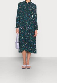 Blommig långärmad klänning i mörkgrön tyg, med en knapp-up framsida och en flödande silhuett. Matchad med en lavendelfärgad handväska och sandaler.