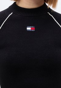 Μαύρο πλεκτό μπλουζάκι με ριγέ υφή και ψηλό γιακά, με μικρό κεντητό λογότυπο Tommy Jeans στο στήθος και λευκή ραφή ως διακόσμηση.