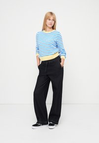 Sinivalkoraidallinen neule keltaisilla reunoilla, yhdistettynä löysiin mustiin housuihin ja mustiin lenkkareihin. Malli seisoo käsineen taskuissa.