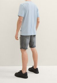 Una persona está de espaldas al espectador, vistiendo una camiseta de color azul claro y pantalones cortos grises, combinados con zapatillas negras. El fondo es sencillo.