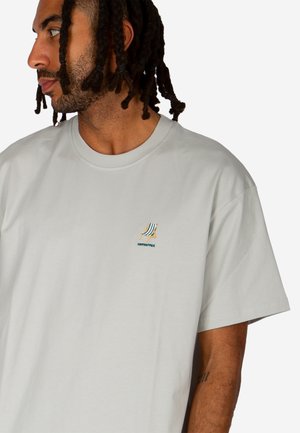 Uomo con capelli intrecciati che indossa una maglietta grigio chiaro con una piccola sedia a sdraio a righe ricamata e testo sul petto sinistro.