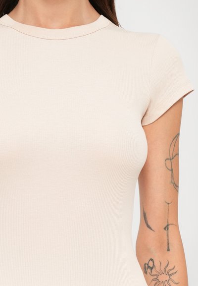 Primo piano di una persona che indossa una maglietta a maniche corte a coste di un leggero beige, con tatuaggi sul braccio destro superiore.