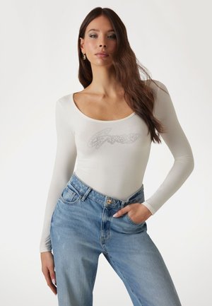 Bodysuit blanco de manga larga con un logo plateado brillante, combinado con jeans de tiro alto en azul claro. Tejido suave y diseño ajustado.