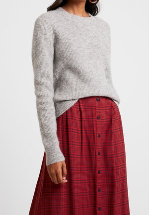 Femme portant un pull en maille gris doux rentré dans une jupe à carreaux rouge et noir avec des manches longues et des boutons sur le devant.