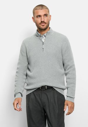 Mann trägt einen hellgrauen Pullover mit Viertel-Reißverschluss über einem gemusterten Hemd und dunkelgraue Hose, blickt leicht nach links.