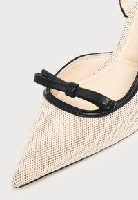 Gros plan d'une chaussure plate beige à bout pointu avec une bordure noire et un petit nœud noir sur le bord supérieur avant.