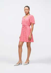 Γυναίκα με πλεξούδες, φοράει ροζ φόρεμα με κοντά μανίκια και λευκές γόβες με λουράκι, στέκεται μπροστά σε απλό λευκό φόντο.