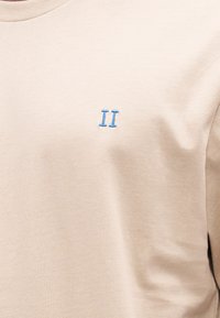 Beżowa bawełniana koszulka z okrągłym dekoltem i krótkimi rękawami. Na piersi niebieskie haftowane logo "II", gładka faktura.