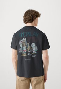 Svart bomulls t-shirt med en färgglad grafisk design av spelkort, tärningar och pokerchips med texten "REPLAY HIGH STAKES" på baksidan.
