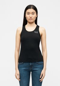 Νεαρή γυναίκα με μακριά σκούρα μαλλιά που φοράει μαύρο αμάνικο τοπ με το λογότυπο "EA7 Emporio Armani" και μπλε τζιν, στέκεται μπροστά σε λευκό φόντο.