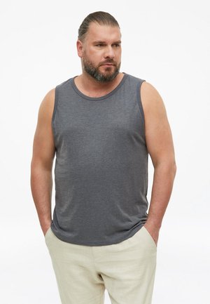 Mann mit Bart und zurückgekämmtem Haar, der ein graues ärmelloses Shirt und helle Hosen trägt, mit den Händen in den Taschen vor weißem Hintergrund.