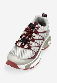 Zapatilla deportiva gris claro y burdeos con parte superior de malla, cordones elásticos tipo toggle, collar acolchado y suela de goma texturizada.