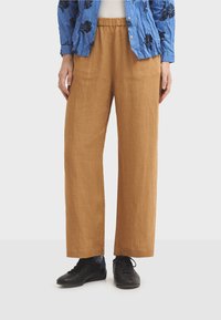 Pantalon ample en lin marron clair avec taille élastique, présentant une texture lisse et une coupe droite. Des chaussures noires complètent la tenue.