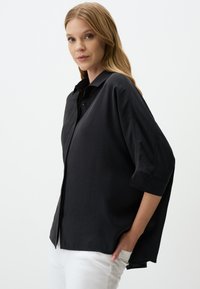 Black button-up shirt with a loose fit, short sleeves, and a soft texture. Features a classic collar and a curved hemline.