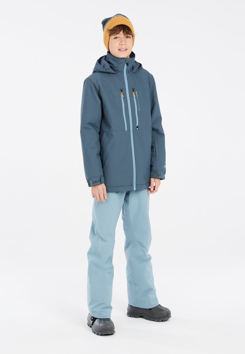 Giacca impermeabile blu con cappuccio, pantaloni azzurri, e stivali neri. Caratterizzata da tasche frontali e cerniere a contrasto. Berretto giallo.