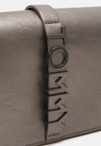 Sac à main en cuir gris texturé avec une sangle verticale ornée de lettres métalliques foncées formant "TOMMY."