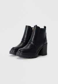 Bottines noires en simili cuir lisse, avec un talon épais, une sangle décorative et une fermeture éclair visible sur le côté.
