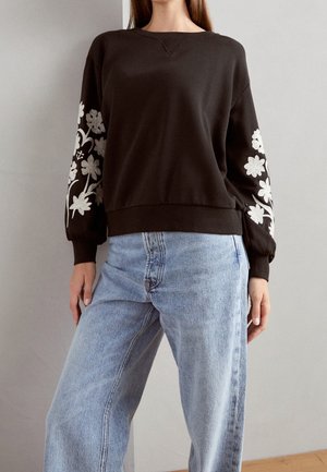 Woman wearing a dark brown sweatshirt with white floral embroidery on sleeves and light blue high-waisted jeans, standing indoors.