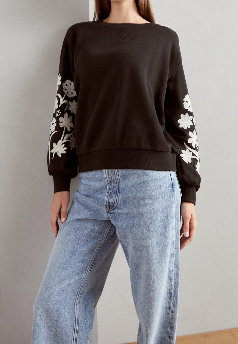 Femme portant un sweat-shirt marron foncé avec une broderie florale blanche sur les manches et un jean bleu clair taille haute, debout à l'intérieur.