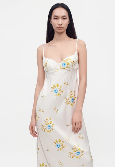 Femme aux longs cheveux noirs portant une robe blanche à fines bretelles avec des motifs floraux bleus et jaunes, debout devant un fond blanc.