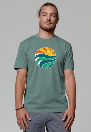Groene katoenen T-shirt met een cirkelvormige grafiek met een gele zon, blauwe golven en groene accenten, weergegeven tegen een lichtgrijze achtergrond.