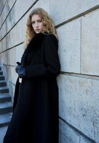 Abrigo de lana negra con cuello de piel, combinado con guantes de cuero negro. El modelo está de pie frente a una pared de piedra, mostrando una silueta elegante.