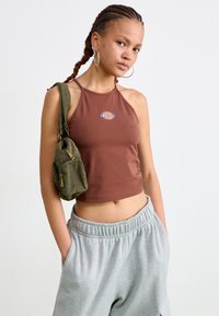 Brun halterneck crop top med brodert logo, kombinert med lysegrå joggebukser og en grønn kordfløyelsveske med glidelås.