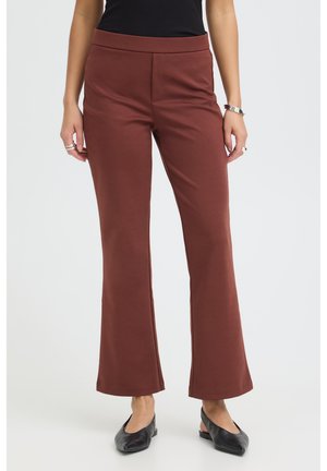 Femme portant un pantalon marron droit, un haut noir, des ballerines noires, des bracelets et des bagues, debout devant un fond clair uni.