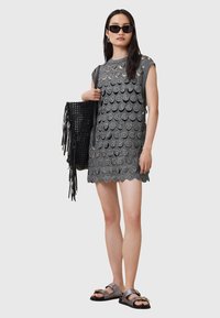 Rochie gri fără mâneci cu un design circular asemănător pălăriilor de paie și un tiv ondulat. Geantă neagră din material țesut cu detalii franjurate. Ochelari de soare negri și sandale.