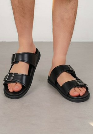 Personne portant des sandales en cuir noir avec deux brides à boucle réglables, debout sur un sol gris devant un fond uni.