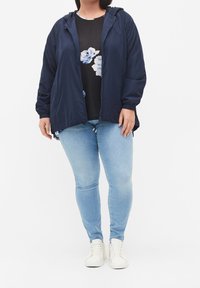 Kvinna iklädd en marinblå zip-up jacka över en svart blommig topp, ljusblå jeans och vita sneakers, stående mot en vit bakgrund.