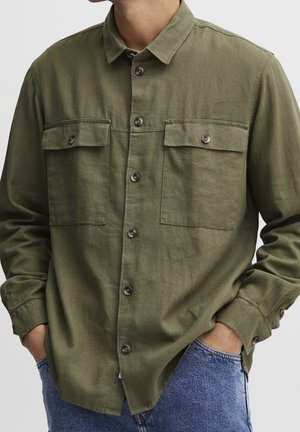Homme portant une chemise boutonnée vert olive avec deux poches poitrine boutonnées et un jean bleu, les mains dans les poches.