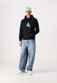 Schwarzer Hoodie mit dreieckigem blauen Grafikdesign, lässige hellblaue Jeans und weiße Sneakers. Das Modell trägt eine hellgraue Mütze und hat die Hände in den Taschen.
