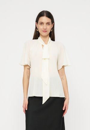 Mujer vestida con una blusa blanca de manga corta con un lazo delantero y detalles plisados, combinada con una falda negra sobre un fondo liso.