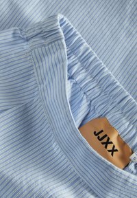 Gaiši zils svītrains audums ar elastīgu jostu; birka ar "JJXX" ir redzama. Gluda struktūra un maiga apdare.