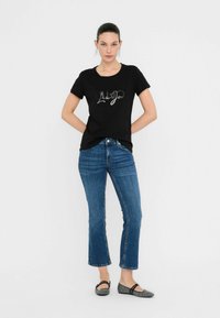 Zwarte T-shirt met korte mouwen met zilveren "Hugo" tekst, gecombineerd met blauwe jeans. Model draagt zwarte en zilveren ballerina's; eenvoudig, casual design.