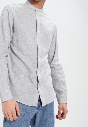 Mężczyzna ubrany w jasnoszarą koszulę na guziki z długim rękawem i niebieskie dżinsy, stojący na tle jednolitego jasnego tła.