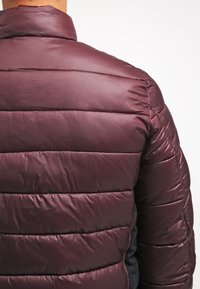 Burgundy quiltad puffjacka med en slät textur och sömdetaljer. Har en hög krage och kontrasterande tyg på sidorna.